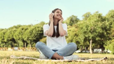 Bir sürü mutlu, güzel, beyaz kadın, kulaklık takıyor, müzik dinliyor, hareket ediyor, yeşil çimlerin üzerinde bacakları bağlı oturuyor, gülümsüyor, el sallıyor ve kameraya bakıyor.