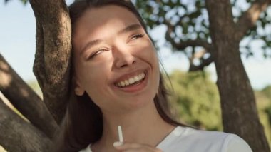 Genç, güzel, beyaz bir kadının plastik kamışla içki içişi, gülümsemesi, güneşli yaz gününde ağaç dalına yaslanışı.
