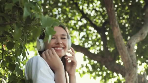低角度胸アップPovの喜び若いです白人女性の耳の上のヘッドフォンを身に着けています 音楽を聴きます 晴れた夏の公園の木の枝の間に立って 笑顔とカメラを見ます — ストック動画