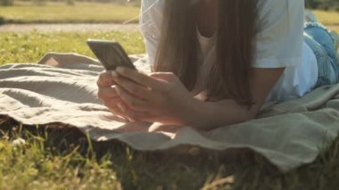 Genç, uzun saçlı, beyaz bir kadının çimlerin üzerinde karnı üzerinde yatması, akıllı telefon kullanması, gülümsemesi, sonra arkasını dönmesi. Güneşli yaz gününde eğlenen bir kadın.