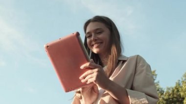 Yeşil ağaçların ve açık havada mavi gökyüzünün önünde gülümseyen, güneşli bir günde pembe kutuda tablet bilgisayar kullanan, daha sonra kameraya bakan genç, keyifli beyaz kadının düşük açılı görüntüsü.