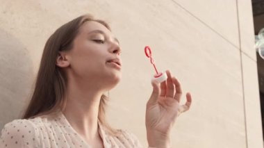 Düşük açılı, mutlu, beyaz bir kadın gülümsüyor, dışarıda sabun köpükleri üflüyor.