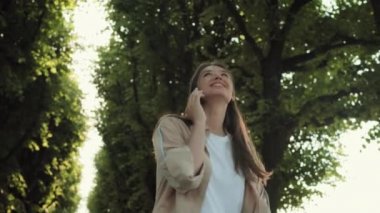 Düşük açılı orta ölçekli mutlu, uzun saçlı beyaz kadın gülümseyen, yürüyen, dönen, cep telefonuyla konuşan güneşli yaz gününde.