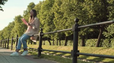 Geniş açıda, mutlu beyaz kadın gülümsüyor, parktaki uzun yeşil ağaçların önünde siyah metal çitlere yaslanıyor, güneşli bir günde pembe kutuda tablet bilgisayar kullanıyor.