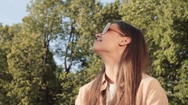 Genç, mutlu, beyaz bir kadının gülümseyen, güneşli yaz parkında oturan, güneş gözlüğünü çıkaran, parmaklarını şaklatan, işaret eden, göz kırpan ve kameraya bakan görüntüsü.