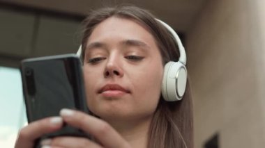 Kablosuz kulaklık takan, akıllı telefon kullanan ve kameraya bakıp gülümseyen neşeli, beyaz bir kadının düşük açılı görüntüsü.