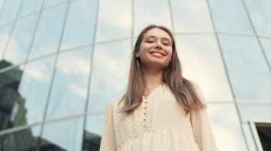Düşük açılı, kahverengi saçlı, heyecanlı, beyaz kadın camdan binaya bakıyor, sonra başını çeviriyor, gülümsüyor, kameraya el sallıyor.