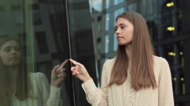 Orta ölçekli, kahverengi saçlı, heyecanlı beyaz bir kadının yürüdüğü, modern binanın cam cephesinde parmaklarını gezdirdiği, sonra yukarı bakıp kameraya gülümsediği bir görüntü.