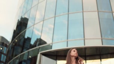 Çerçeveye giren, kağıt alışveriş torbaları tutan, görünmez arkadaşı gören, el sallayan, gülümseyen ve ayrılan genç beyaz kadının düşük açılı görüntüsü.