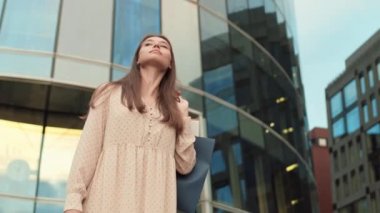 Düşük açılı, neşeli, beyaz bir kadın. Cam binanın önünde duruyor, elinde poşetler, görünmez bir arkadaş görüyor, el sallıyor, gülümsüyor ve gidiyor.