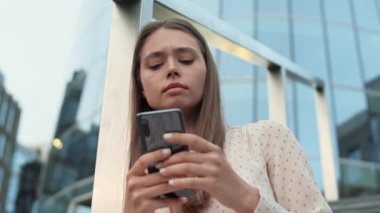 Düşük açılı göğüs hizasında, kahverengi saçlı beyaz kadın akıllı telefon kullanıyor, metal çitlere yaslanıyor, etrafa bakıyor.