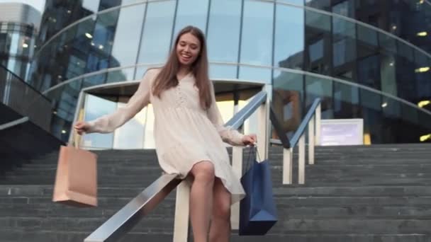 一群年轻的棕色头发的高加索女人 面带微笑 穿着夏装 手里拿着购物袋 从现代建筑外的楼梯金属扶手上滑落下来 — 图库视频影像