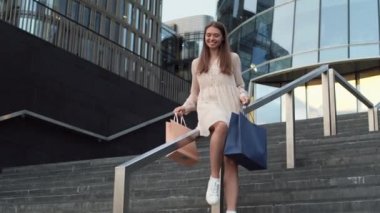 Genç, kahverengi saçlı, beyaz bir kadın. Yazlık elbise giyiyor. Elinde poşetlerle modern binaların dışındaki metal merdivenlerden kayıyor.