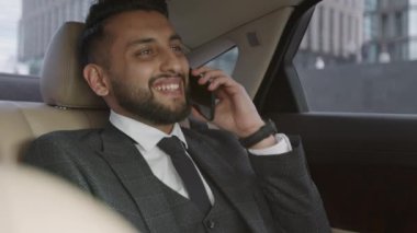 Gülümseyen, gri takım elbiseli, cep telefonuyla konuşan, arabanın arka koltuğunda oturan, sonra da kameraya bakan neşeli, genç, melez iş adamının göğsü kabarmış görüntüsü.