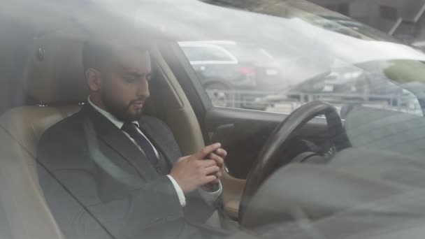 混血青年商人身穿正装 坐在方向盘后面 用手机打电话 从前窗往外看中型轿车 — 图库视频影像