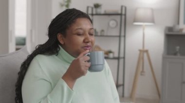 Orta boy bir Afro-Amerikan kadın resmi. Günlük giysiler giymiş, elinde çay fincanıyla evdeki kanepede oturuyor ve tadını çıkarıyor.