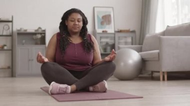 Genç, kıvrımlı, Afro-Amerikan kadın yogilerin spor giysileri giyip kauçuk hasırın üzerinde otururken ve kapalı gözlerle meditasyon yaparken geniş bir açı.