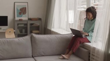 Geniş açıda, sorunlu ciltli ve kafasında dehşet olan genç bir Afro-Amerikalı kadın var. Pencere kenarında oturuyor, koltukta oturuyor ve bilgisayarla çalışıyor.