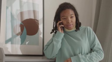 Orta boy bir Afro-Amerikalı kadın resmi, şık saç stili, günlük kıyafetler giymiş, güzel bir resmin önünde duruyor ve telefon görüşmesi yapıyor.