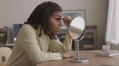 Orta boy Afro-Amerikalı bir kadın. Cildi sorunlu ve dehşetli. Masa aynasının önünde oturmuş kirpiklerine rimel sürüyor.