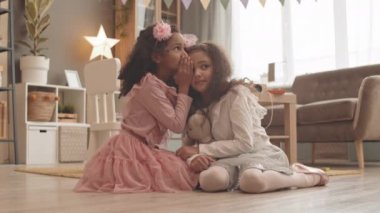 Güzel kıyafetler giymiş, salonda oturmuş dedikodu yapan ve gülümseyen iki sevimli melez kızın tam karesi.