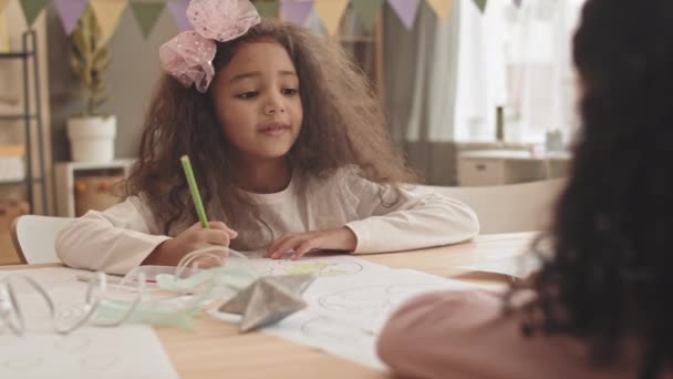 Chest-up malé smíšené-závod dívka v růžové čelenku s lukem, sedí u stolu doma, malování obrázek s tužkou, mluvit s nepoznatelným přítelem před sebou, pak vzít hračku hůlku