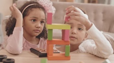 İki sevimli melez kız kardeşin omuz omuza oturması masa başında oturması, renkli ahşap tuğlalarla oyuncak kule yapması, inşaatın kızları korkutması.