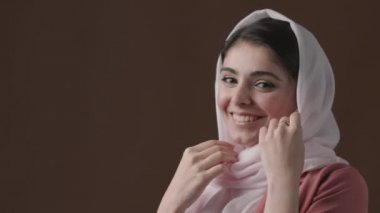 Beyaz başörtüsü ve pembe ceket giyen gülümseyen Müslüman kadının orta boy portresi kahverengi arka planda durup kameraya bakıyor.