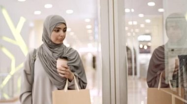 Gri abaya ve tesettürlü genç bir kadının alışveriş merkezine giderken elinde kahve poşeti, elinde kahve fincanı, vitrinin yanında durup ona bakması.