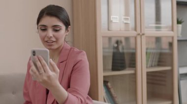 Orta yaşlı, melez bir iş kadınının gündelik kıyafetlerini giyip koltukta elinde telefonla oturduğu ve video bağlantısıyla iş arkadaşıyla video görüşmesi yaptığı bir ortam.