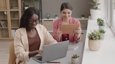 Genç Afrikalı iş kadınının masa başında oturup dizüstü bilgisayarla çalışırken, melez meslektaşının yanında oturup dijital tablet kullanması.