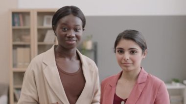 Genç, melez iş kadını ve sıradan kıyafetler giyen Afro-Amerikan iş kadınının ofiste kameraya bakarken çekilmiş bir portresi.