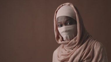 Yüzü kapalı, pembe tesettürlü, kahverengi arka planda duran ve kameraya bakan Müslüman Afrikalı kadının orta boy portresi.