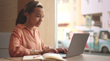 Orta halli, karışık ırktan, modaya uygun kıyafetler ve aksesuarlar giyen, kafeterya masasında oturan, dizüstü bilgisayarla çalışan ve not defterine notlar alan genç bir kadın.