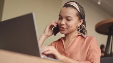 Restoran masasında oturan, telefonla konuşan ve dizüstü bilgisayarla çalışmaya devam eden, modaya uygun giysiler ve aksesuarlar giyen güzel, melez bir kadının düşük açılı görüntüsü.
