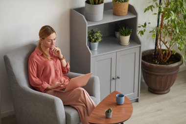 Minimal high angle portrait of modern successful businesswoman using laptop and working while sitting in armchair in stylish home interior, copy space