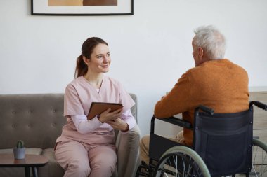 Portrait of smiling female nurse talking to senior man in wheelchair and using digital tablet at retirement home, copy space