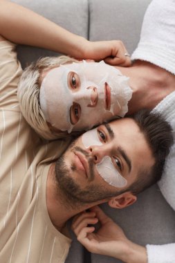 Kanepede yan yana yatıp maske, güzellik ve cilt bakımı yaparken kameraya bakan genç eşcinsel çiftin üst görüntüsü.
