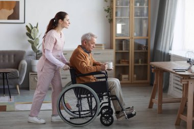 Full length side view portrait of senior man in wheelchair with nurse assisting him at retirement home, copy space
