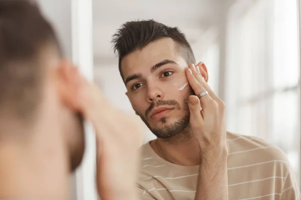 Retrato Hombre Joven Que Aplica Crema Facial Mientras Mira Espejo — Foto de Stock