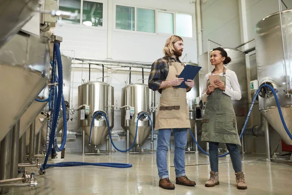 Full length portrait of two young workers wearing aprons looking at each other while standing at brewery workshop, copy space