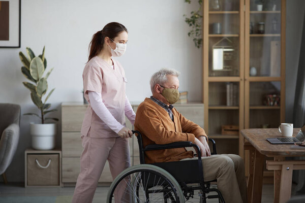 Side view portrait of young female nurse assisting senior man in wheelchair at retirement home, both wearing masks, copy space
