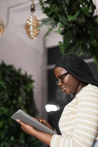 植物によって装飾されたエレガントなオフィスのインテリアに立っている間 デジタルタブレットを使用して若いアフリカ系アメリカ人女性の垂直側のビューの肖像画 コピースペース — ストック写真