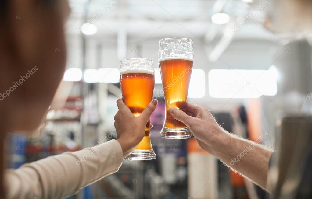 Close up of two workers holding beer glasses while inspecting quality of production and filtration at brewery, copy space