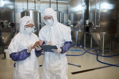 Kimya fabrikasında dijital tablet kullanırken tehlikeli madde giysileri giyen iki genç işçinin belini kaldırın.