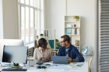 Modern beyaz ofiste masa başında oturmuş projeyi tartışan gülümseyen iki iş adamının yan görünüm portresi.