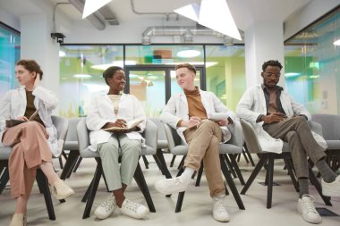 Laboratuvar önlüğü giyen ve üniversitede tıp dersi dinleyen çok ırklı gülümseyen insanların tam boy portresi.