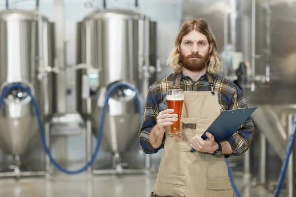 ビールグラスを持ち 醸造工場でワークショップに立っている間にカメラを見ているひげを生やした醸造長の腰の肖像画 コピースペース — ストック写真