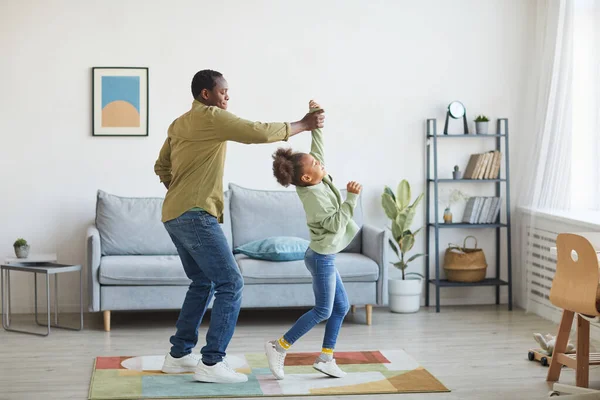 幸せなアフリカ系アメリカ人の父親の完全な長さの肖像画は娘と一緒に踊りながら 最小限の家庭内で楽しみ スペースをコピーします — ストック写真