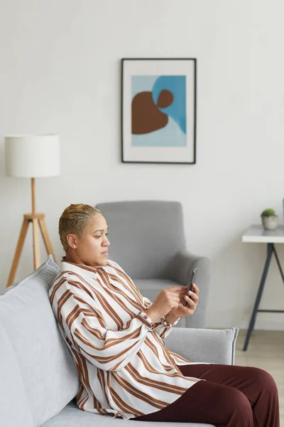 家の中で最小限のソファに座っている間 スマートフォンを使用して現代の混合レース女性の垂直側のビューの肖像画 — ストック写真